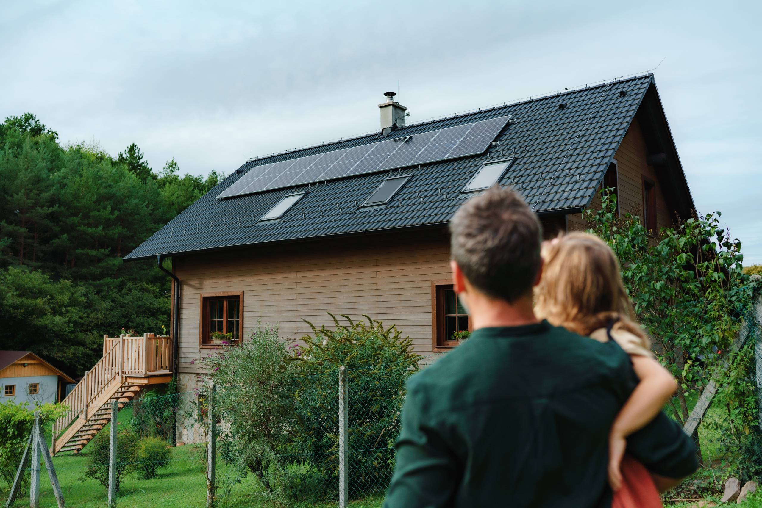 Rear view of dad holding her little girl in arms and looking at their house with solar panels.Alternative energy, saving resources and sustainable lifestyle concept.